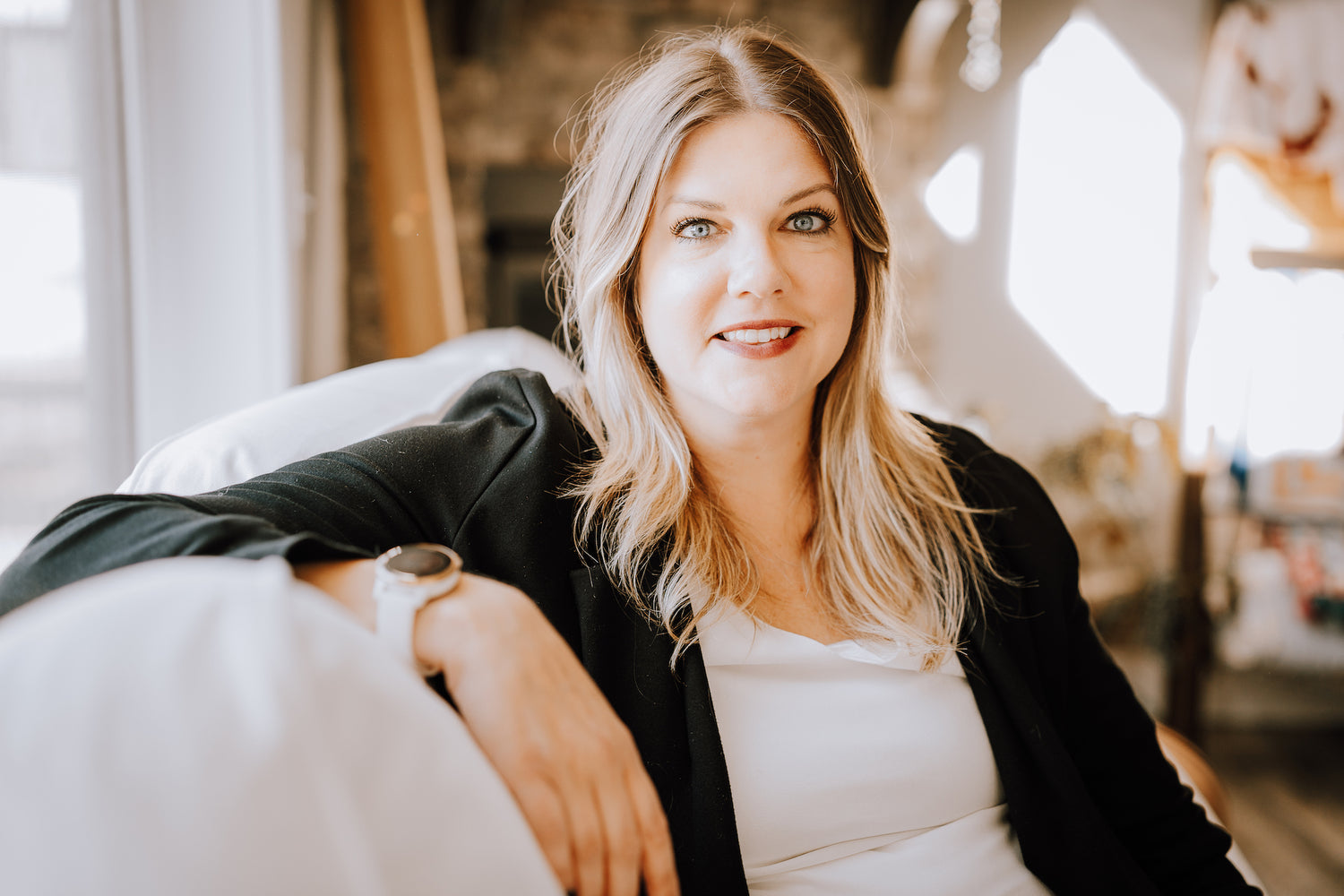 A white woman with blonde hair wearing a black suit jacket over a white shirt sits comfortably on a white couch while smiling at the camera.