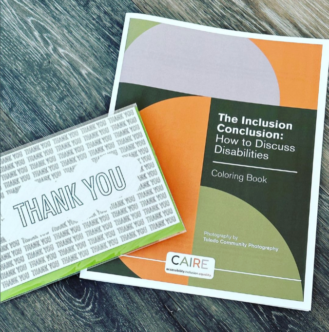 The green, orange, and grey cover of the coloring book (letter sized) called "The Inclusion Conclusion: How to Discuss Disabilities" is laying flat on a table with a thank you card angled on top of it.